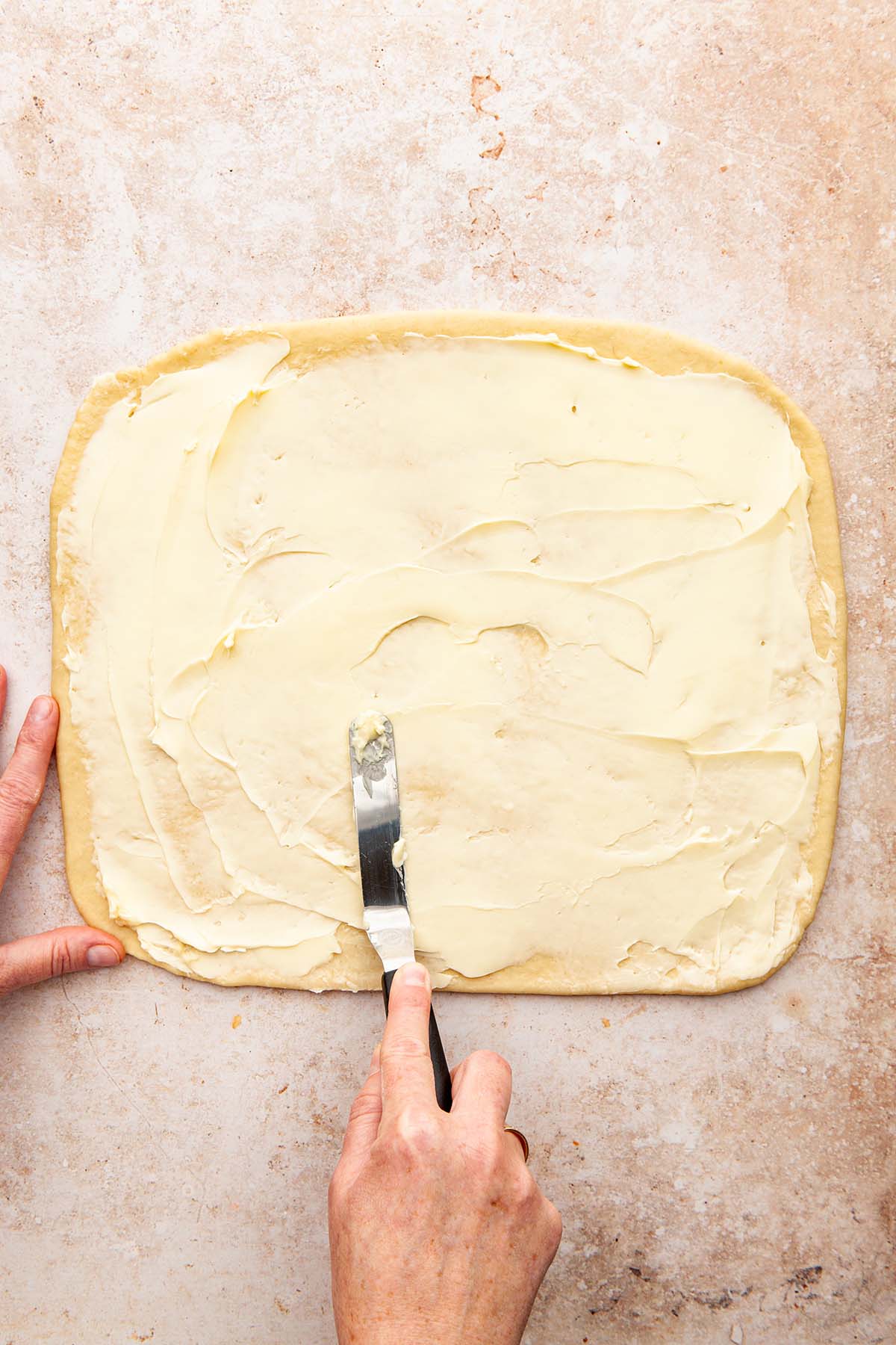 Hands spreading butter onto a rectangle of rolled out dough with an offset spatula.