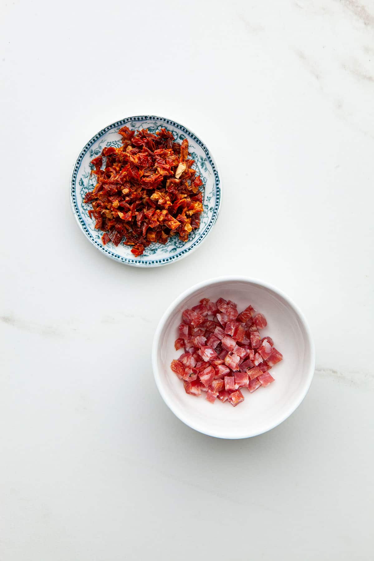 A small dish of finely diced sun dried tomatoes above a small bowl of finely diced Genoa salami.
