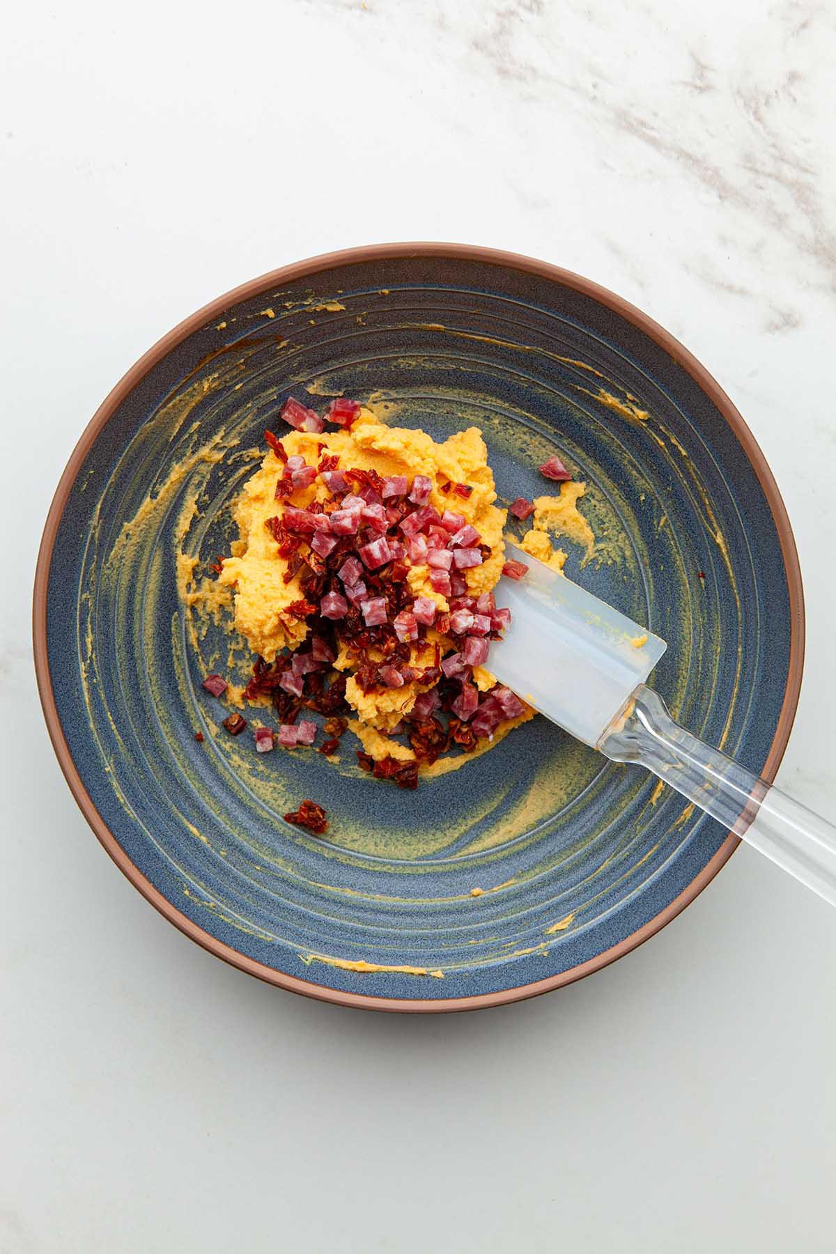 A bowl of mashed egg yolks topped with finely diced salami and finely diced sun dried tomatoes.