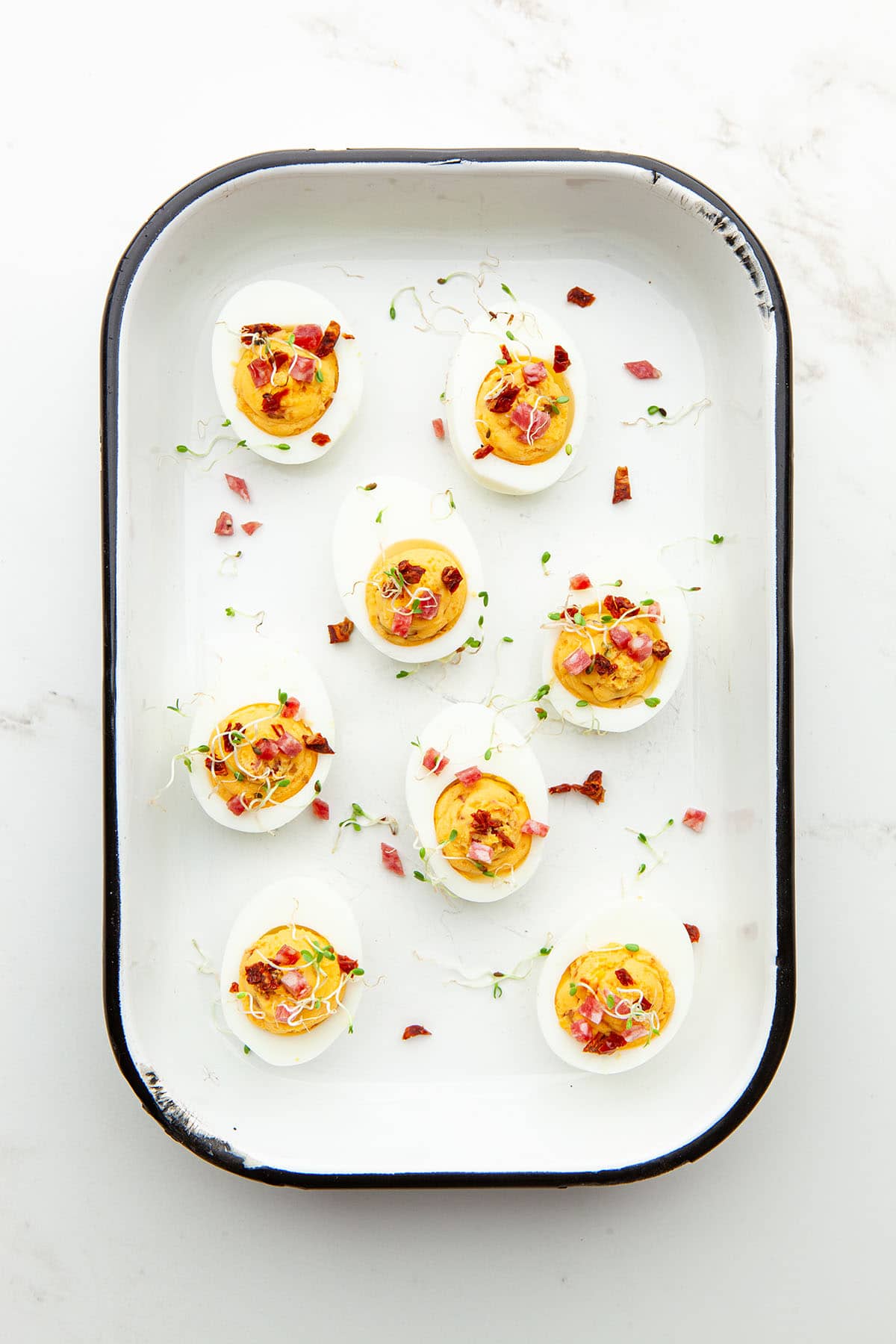 A white enamel tray with black trim and eight sriracha deviled eggs inside garnished with sprouts.