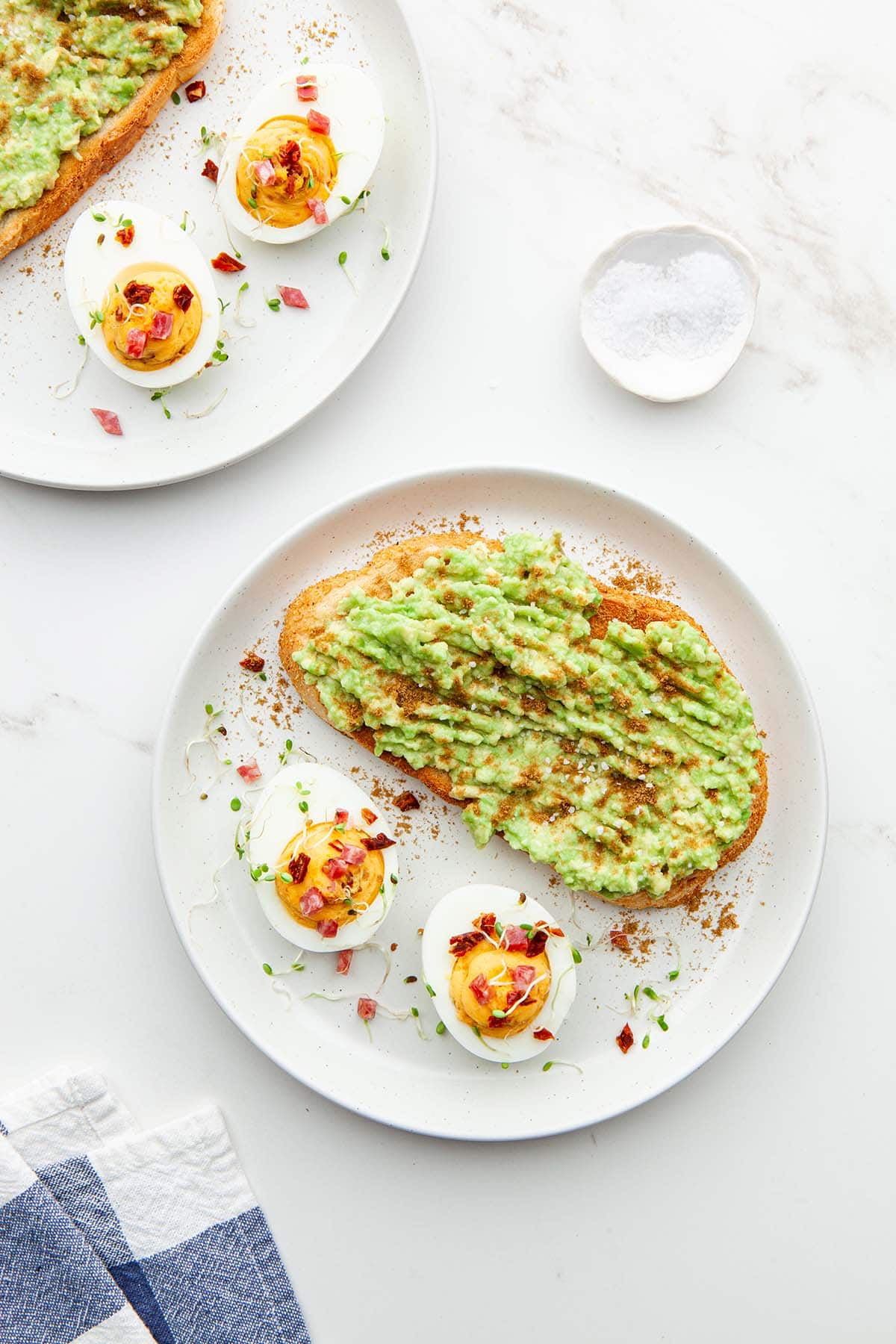 Two plate of avocado toast, each with two sriracha deviled eggs, with a small organically-shaped porcelain dish of coarse salt nearby.