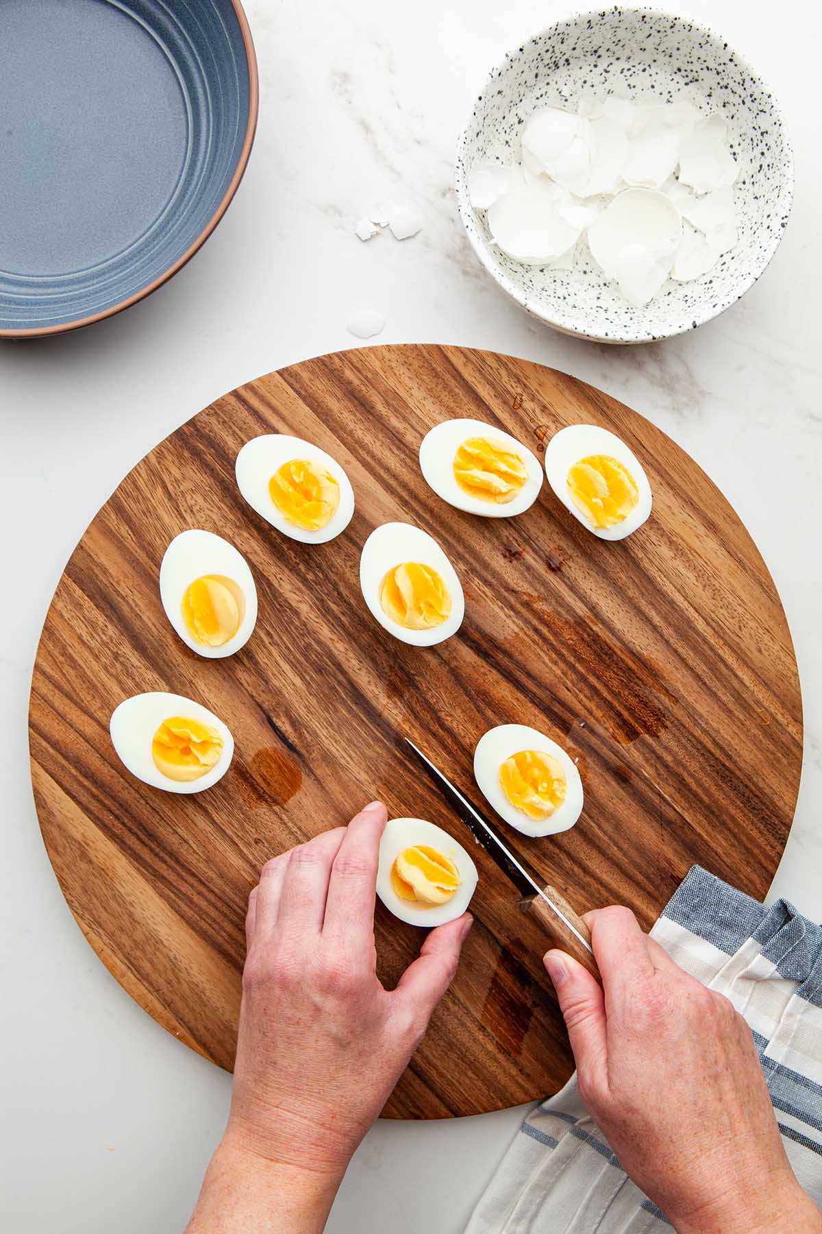 Hands slicing through a hard boiled egg with a paring knife on a wooden cutting board.