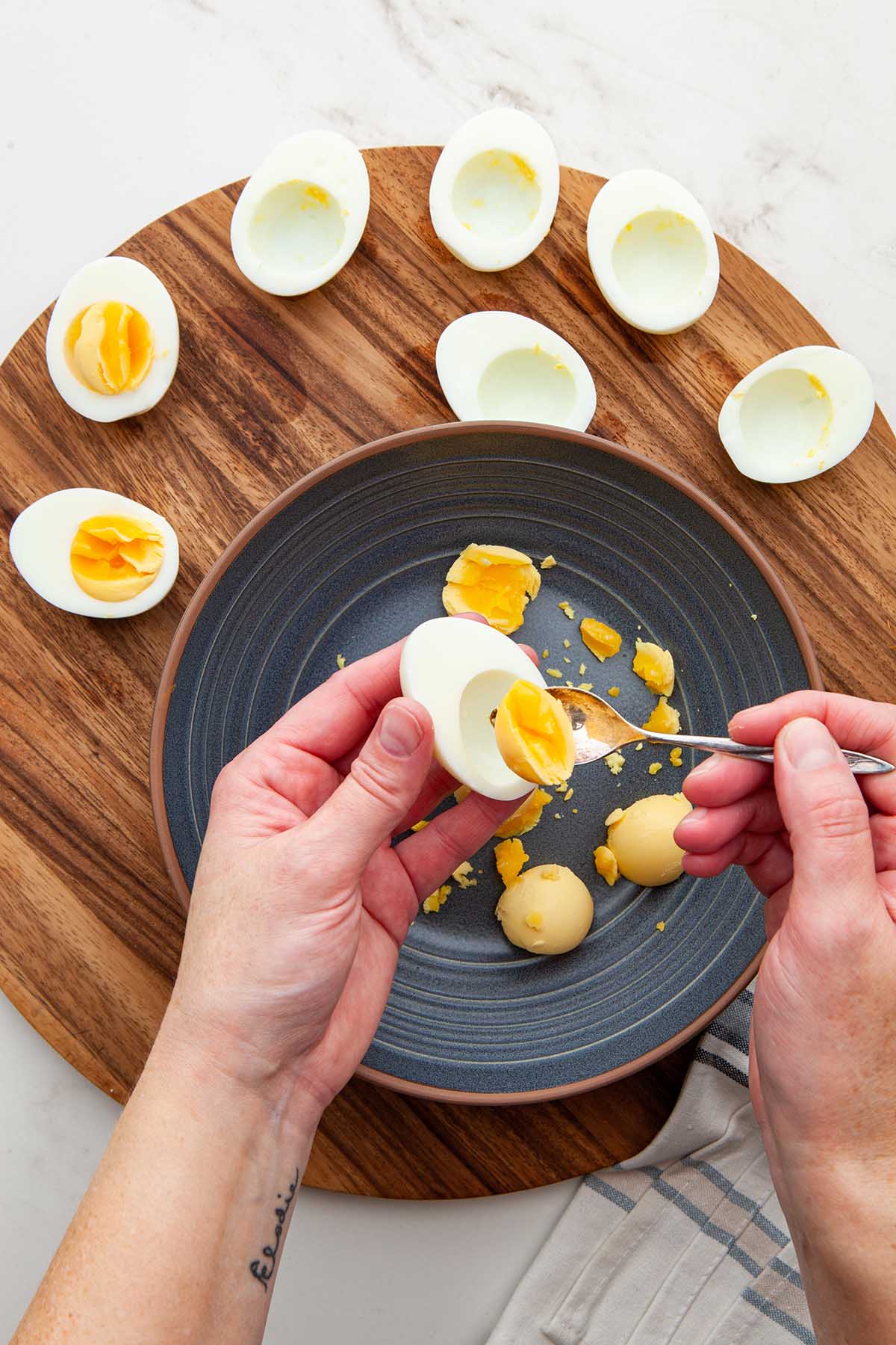 Hands scooping the yolk from a hard boiled egg into a blue bowl on a wooden board.