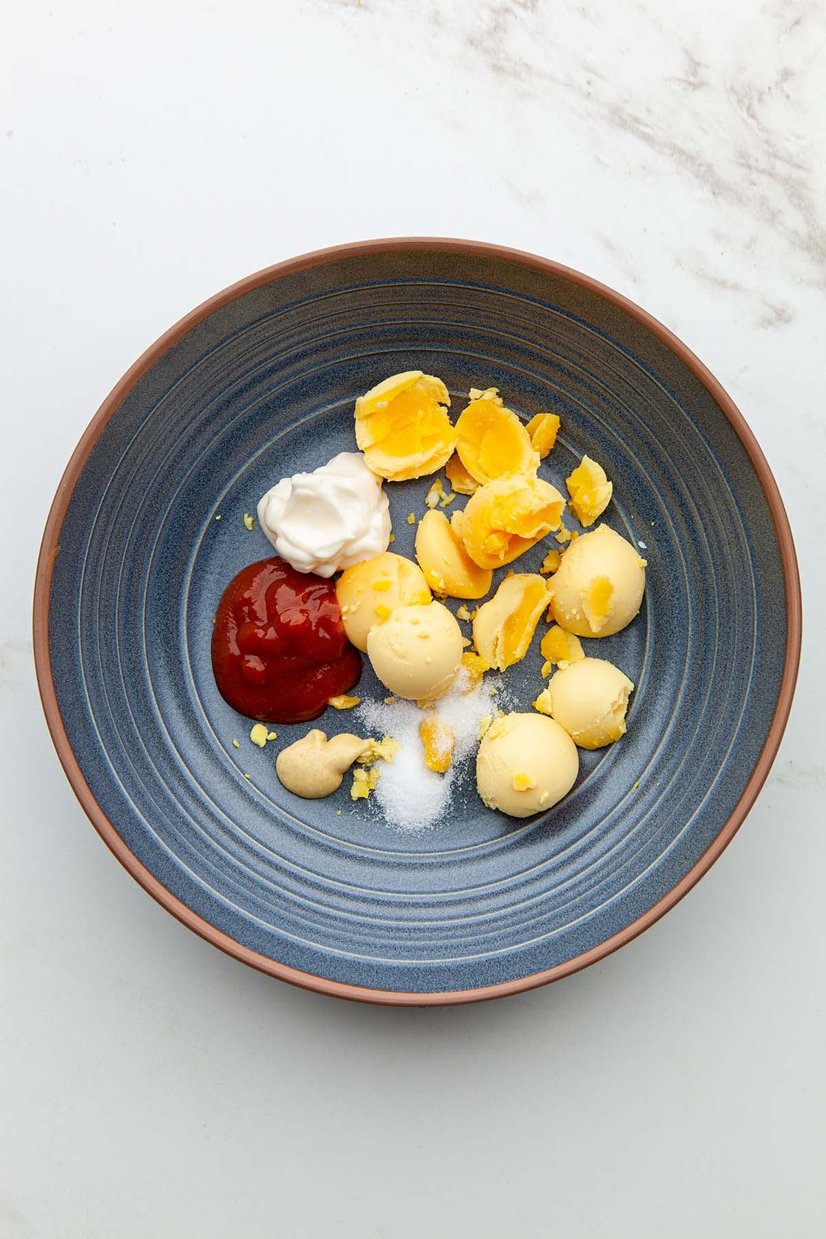 The yolks of hard boiled eggs with sriracha, mayonnaise, Dijon mustard, sugar, and salt in a blue bowl.