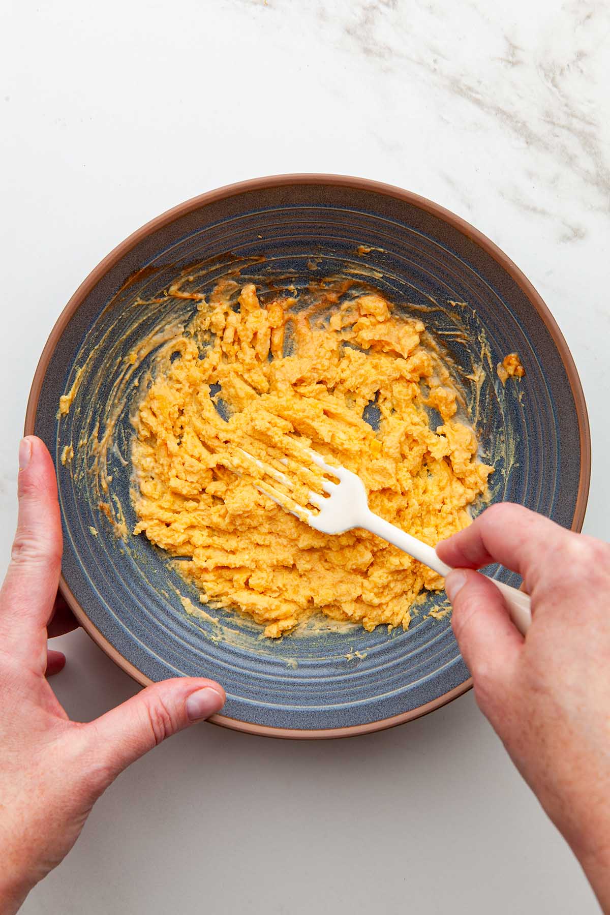 Hands mashing hard boiled egg yolks with sriracha and mayonnaise in a blue bowl using a white fork.
