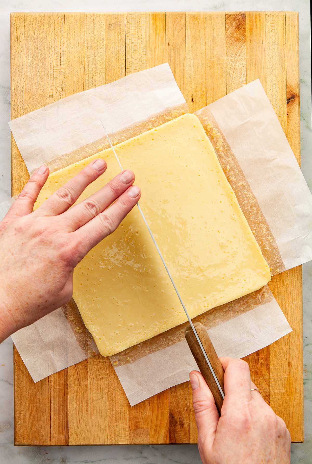 Hands using a long knife to cut lemon bars on a wooden board.