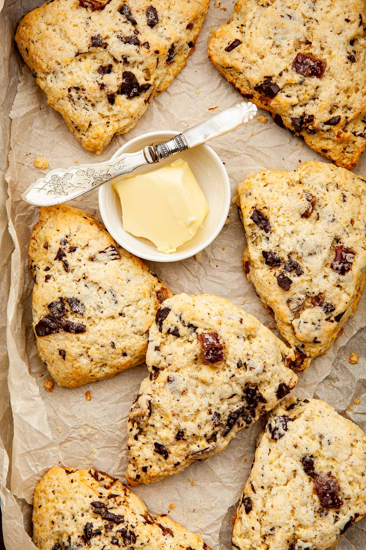 Date and orange scones piled in a box lined with parchment paper with a small dish of butter and a tiny pearl-handled knife.