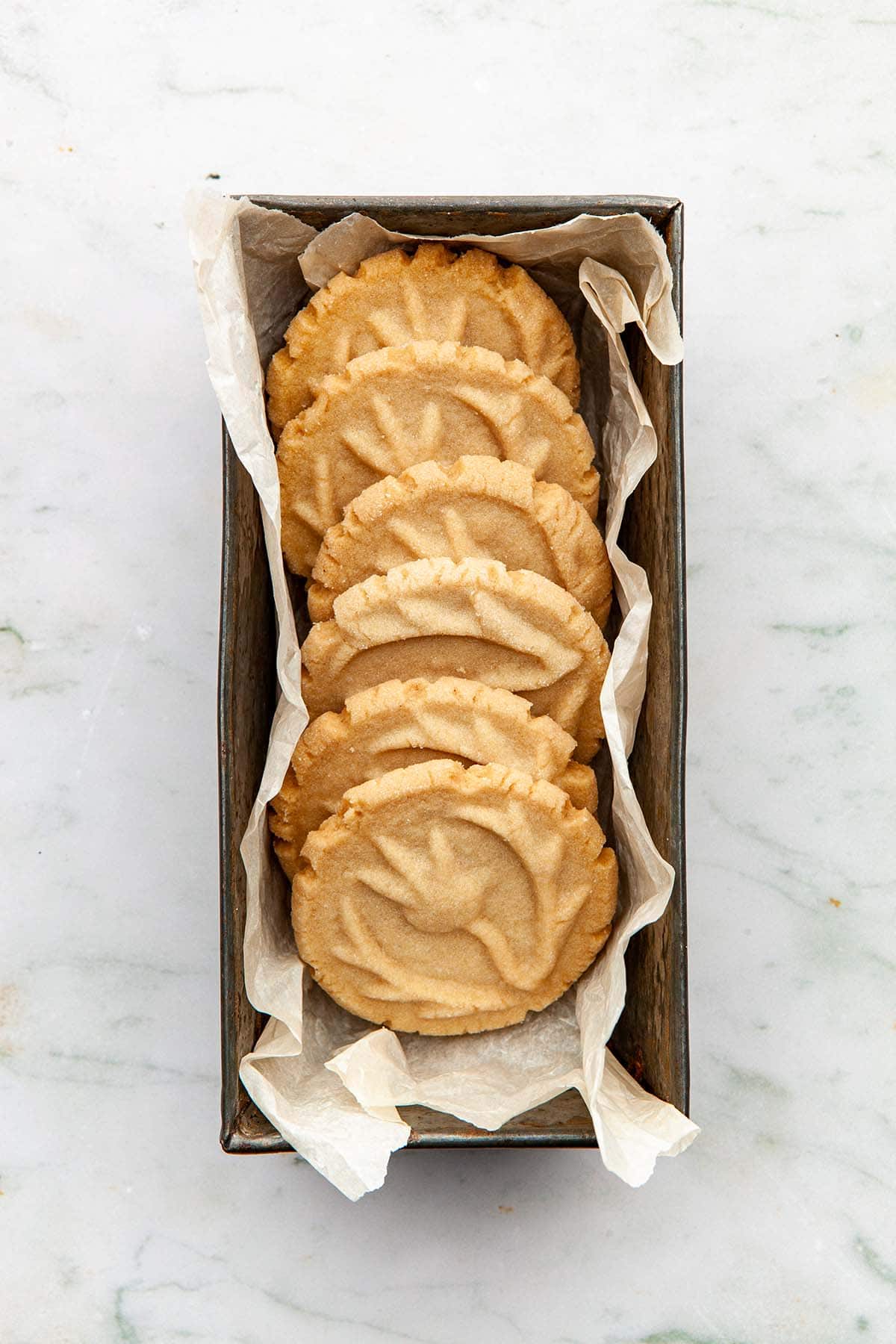 Stamped brown sugar shortbread cookies laying in a small rectangle tin lined with parchment paper.