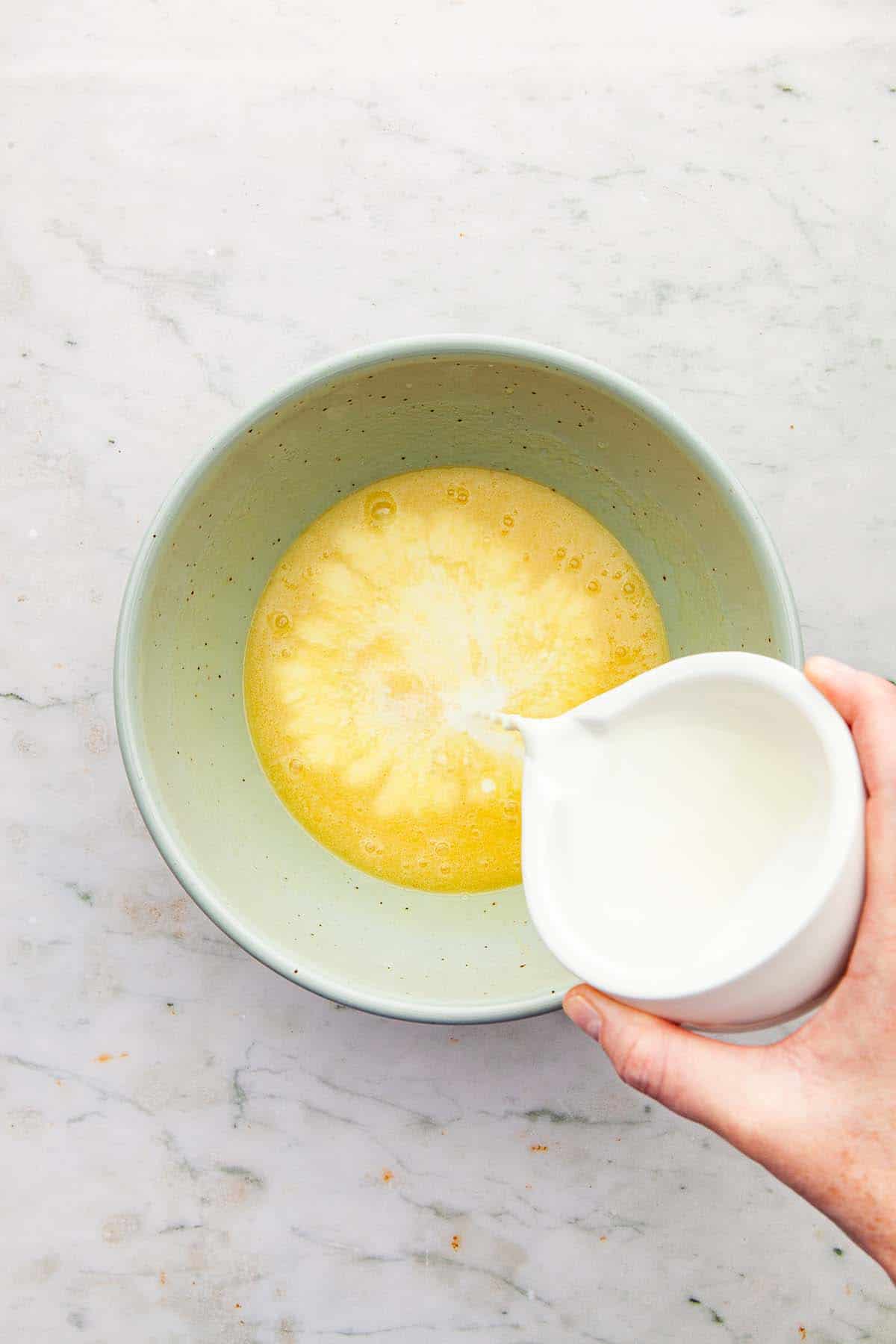 A hand pouring milk into a bowl of whisked eggs and butter from a small white vessel with a spout.