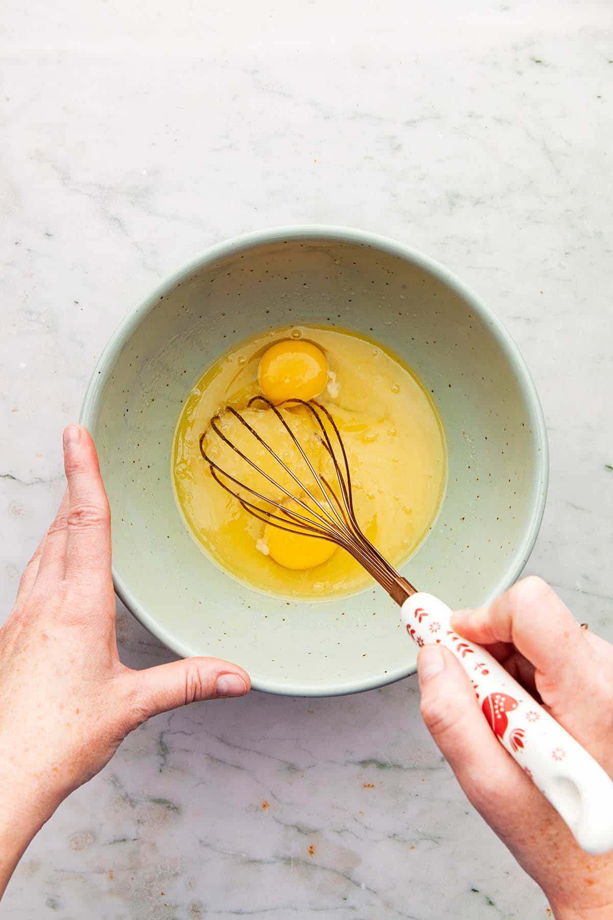 Hands whisking eggs into a bowl of melted butter mixed with sugar.