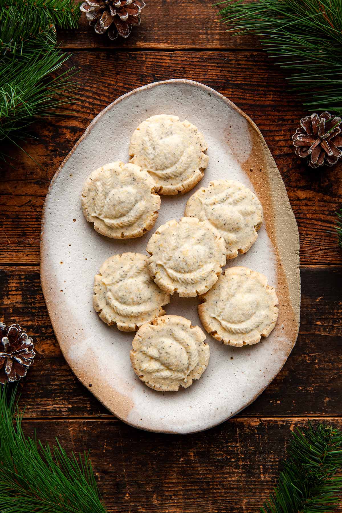 A small platter of lemon poppy seed shortbread cookies surrounded by evergreen branches and a few pine cones.