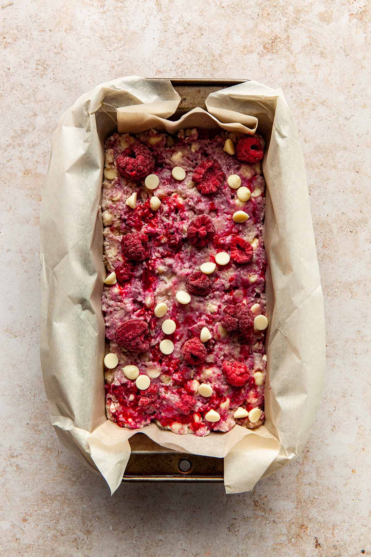 Unbaked batter in a loaf tin lined with parchment paper, topped with extra raspberries and white chocolate chips.