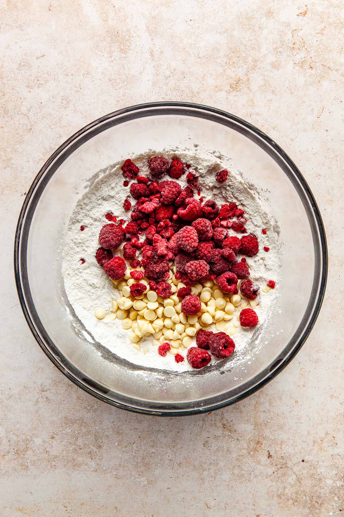 A bowl of dry cake ingredients topped with white chocolate chips and frozen raspberries.