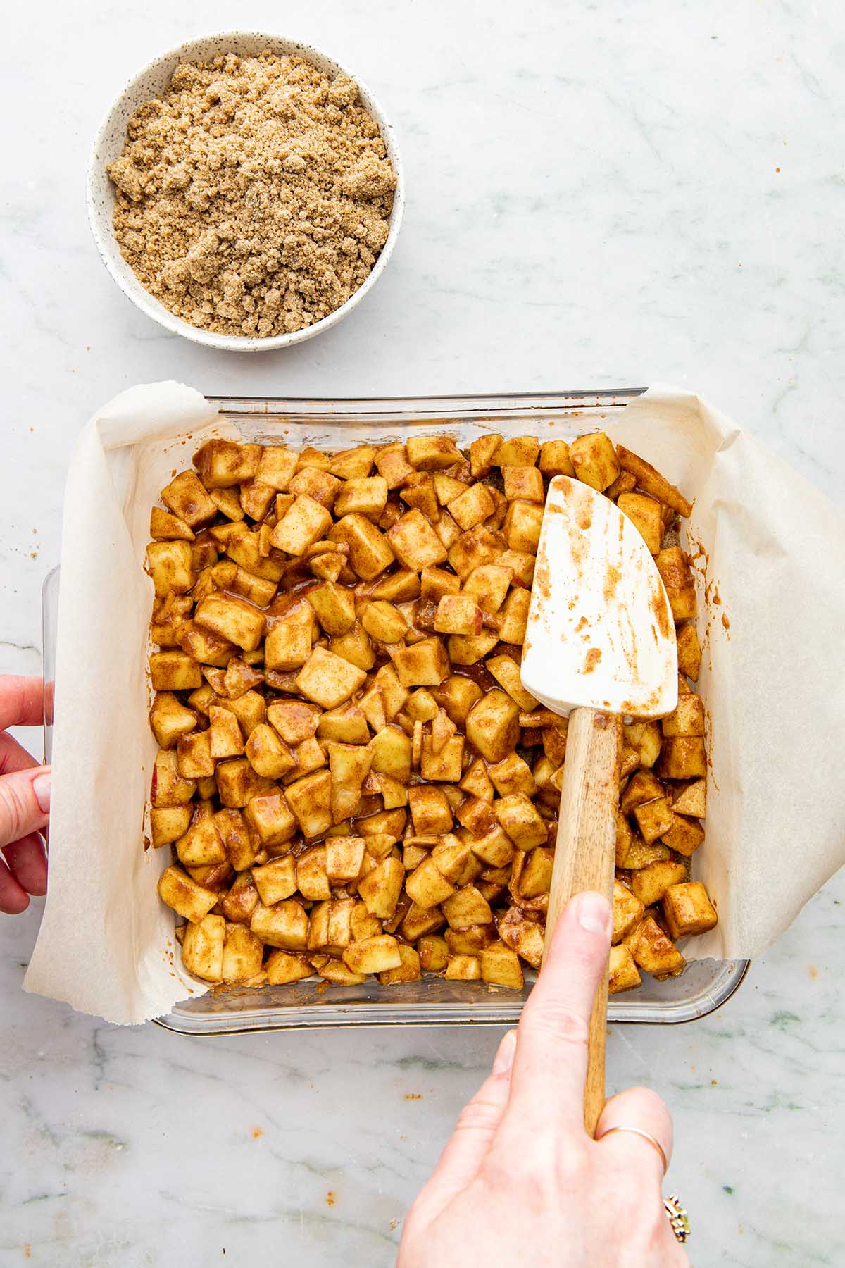 A hand using a rubber spatula to pat chopped apples evenly into a square baking dish.