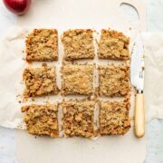 Nine apple crumble bars on a thin plastic cutting board alongside a small knife and some Red Prince apples.