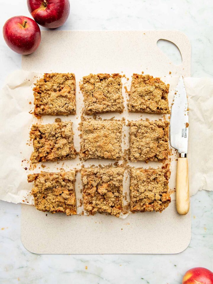 Nine apple crumble bars on a thin plastic cutting board alongside a small knife and some Red Prince apples.