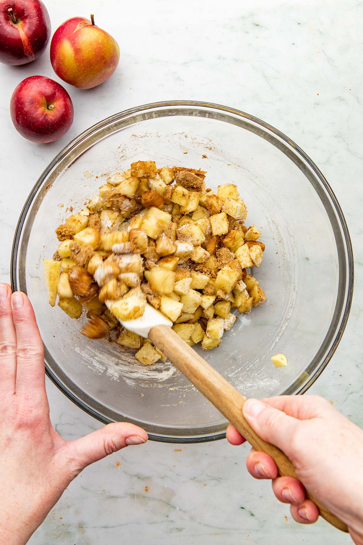 A hand using a rubber spatula to mix chopped apples, cinnamon, brown sugar, rye flour, and salt together in a large glass bowl.