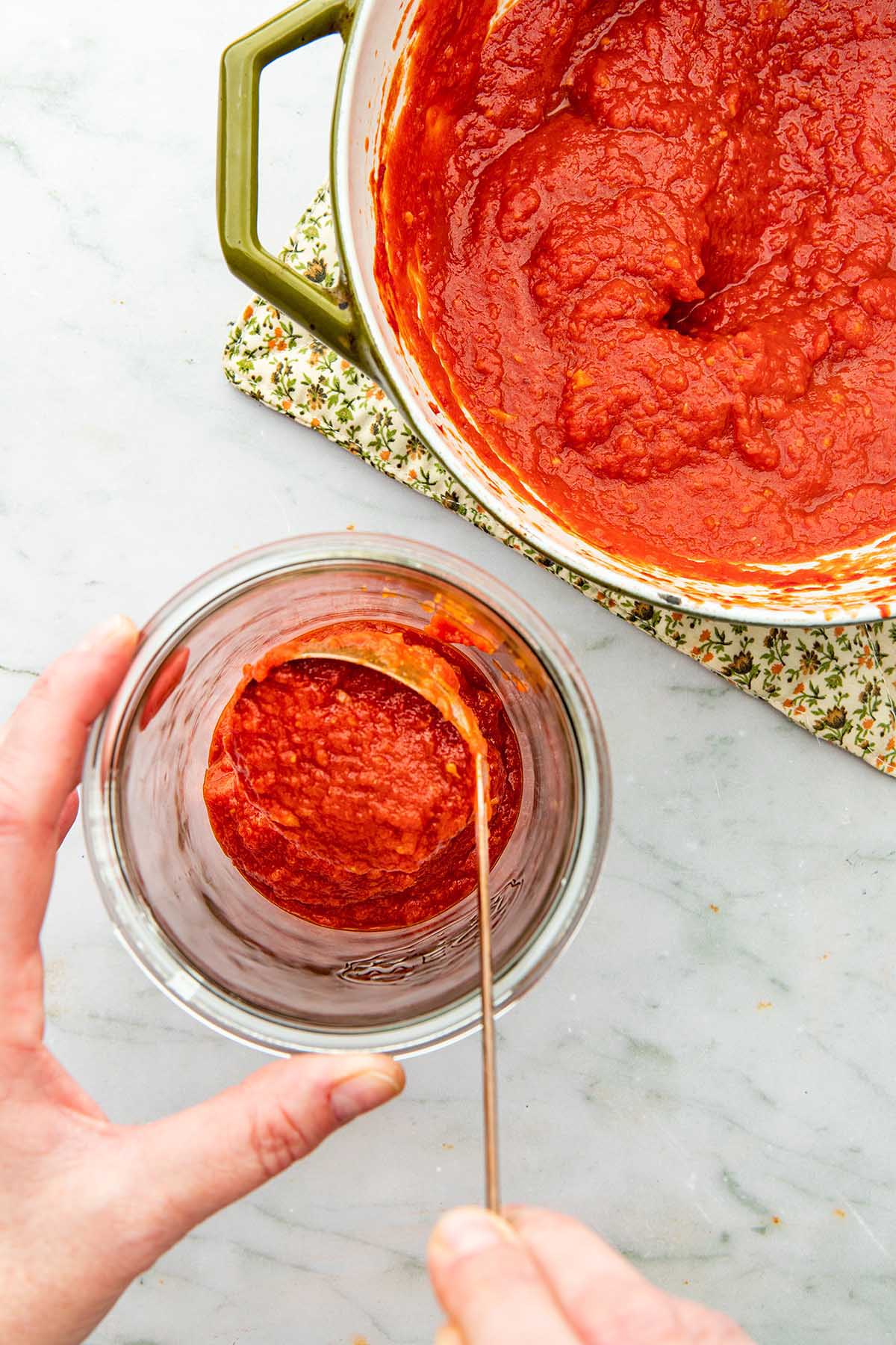 A hand ladling tomato sauce into a glass jar with the pot of cooked sauce nearby.