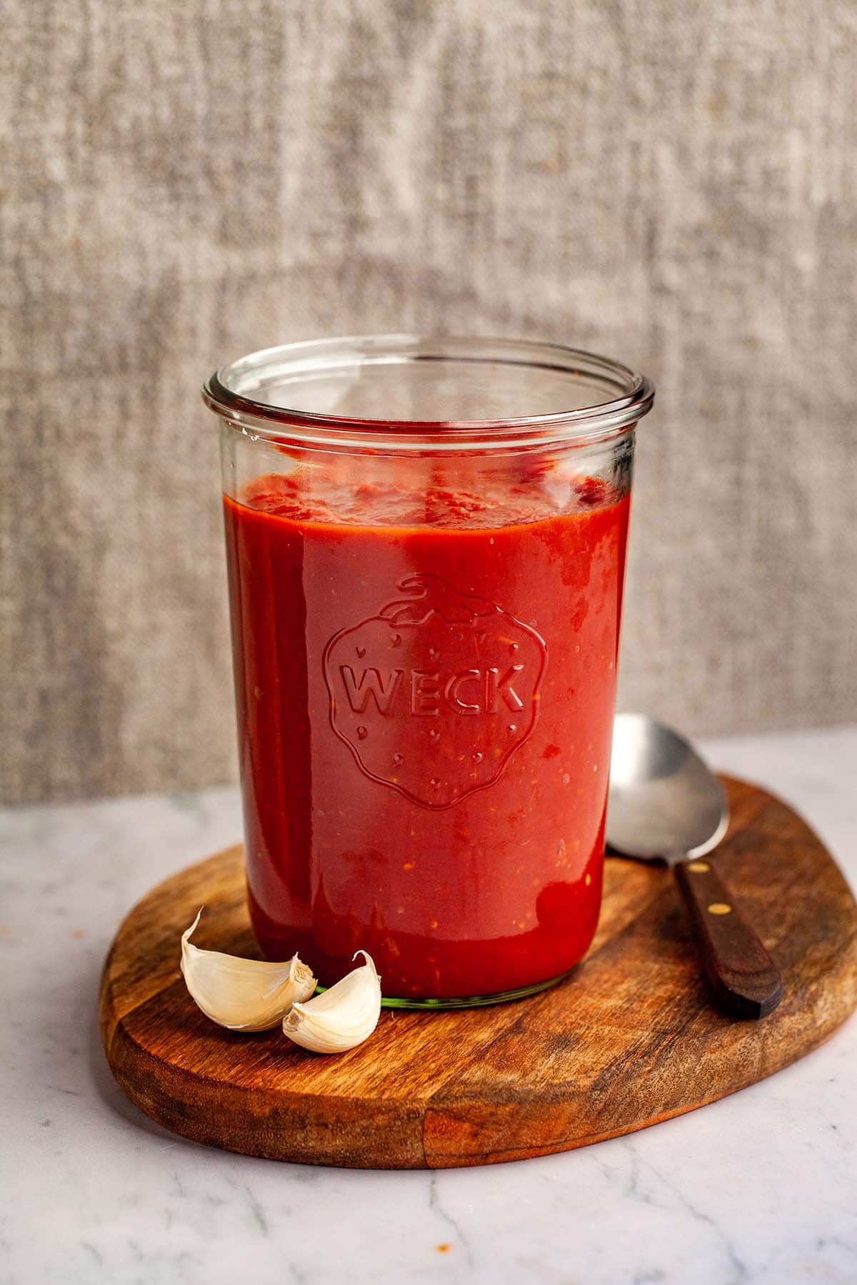 A tall jar of homemade tomato sauce on a wood board with a spoon and two garlic cloves nearby.
