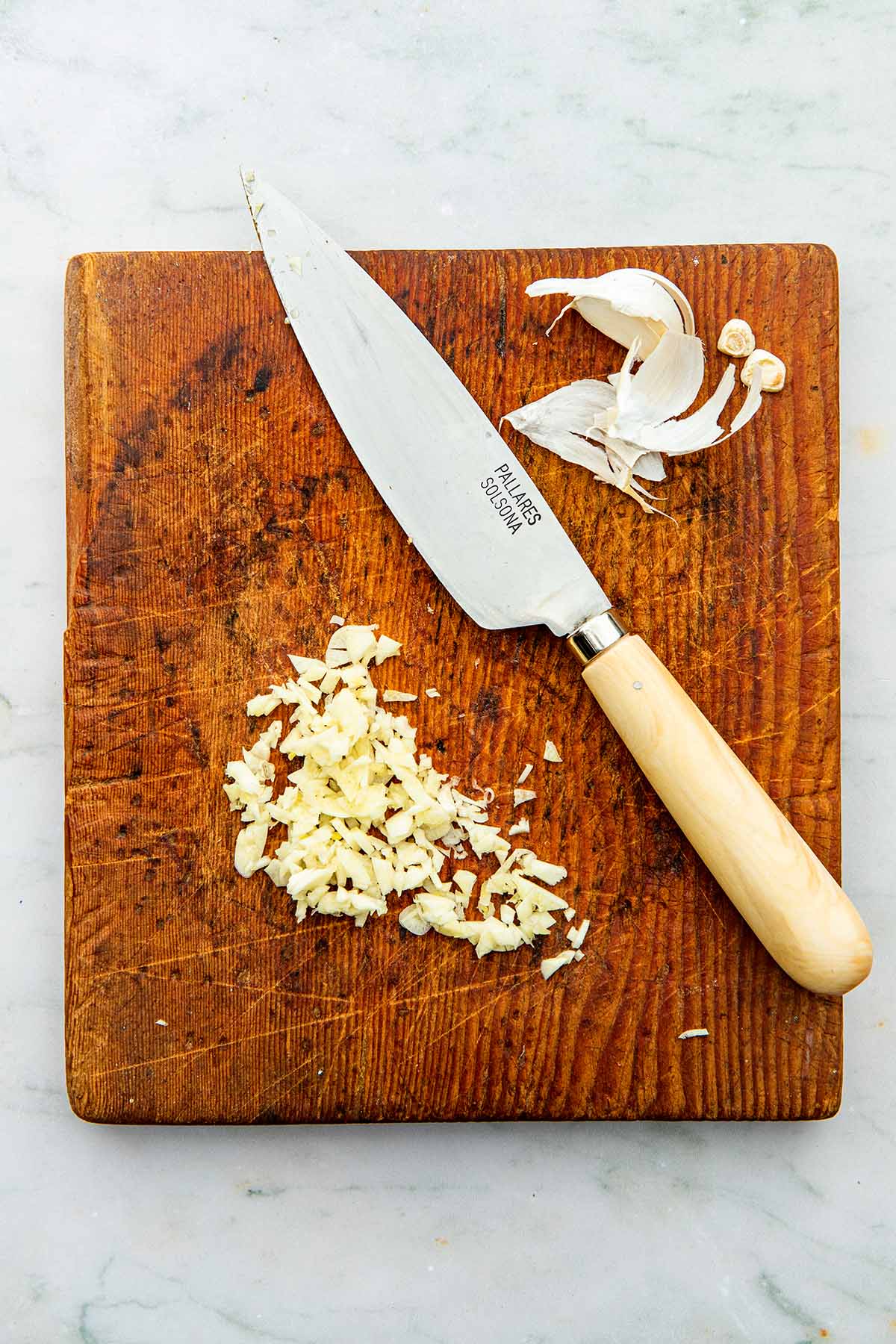A small wood cutting board with chopped garlic, garlic skins, and a knife on top.