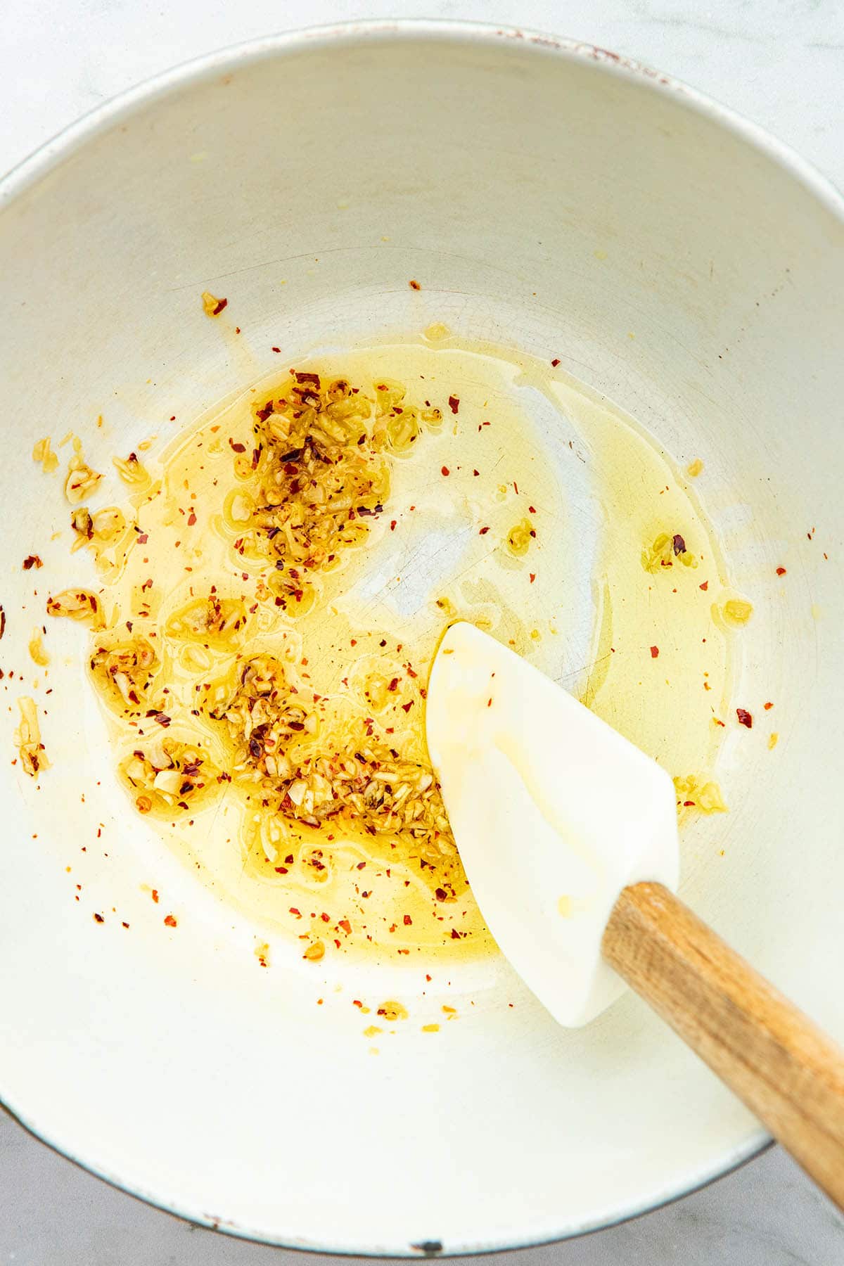 Garlic, olive oil, and red pepper flakes being stirred with a rubber spatula in the bottom of a pot.