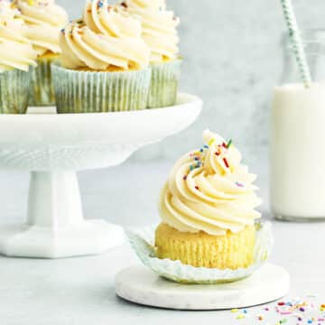 A half-unwrapped cupcake topped with American buttercream and sprinkles. A cake stand with four more cupcakes and a small bottle of milk with a striped straw are nearby