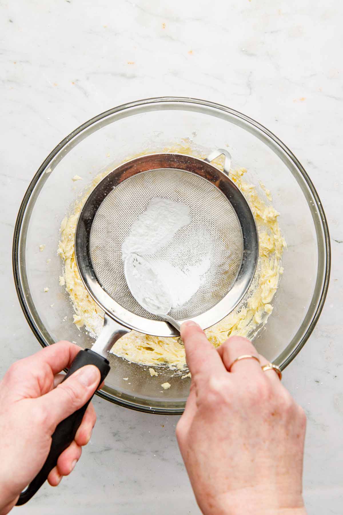 A hand using the back of a spoon to press powdered sugar through a sieve into a bowl of creamed butter.