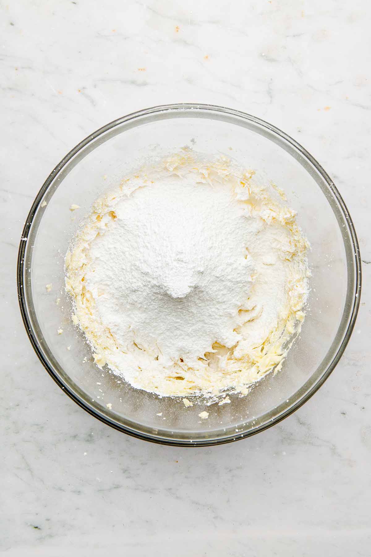 A pile of sieved powdered sugar inside a large glass bowl of whipped butter.