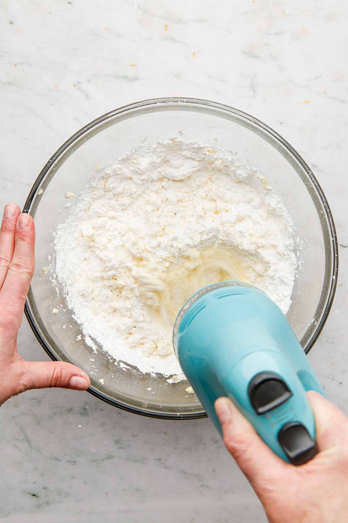 A hand using a hand mixer to mix powdered sugar into creamed butter.