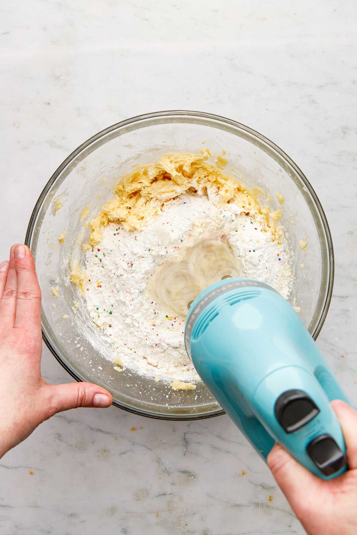 A hand holding a hand mixer to mix flour into cookie dough.