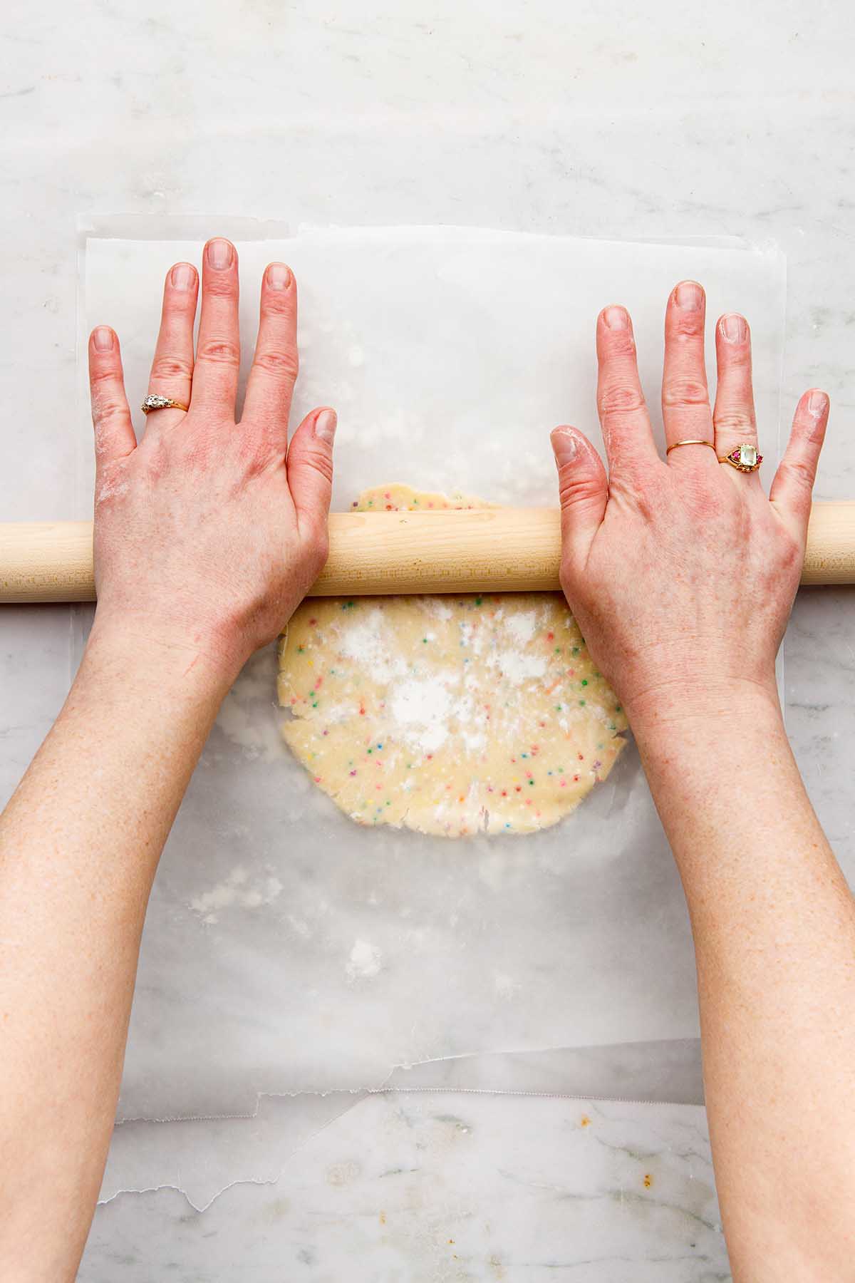 Hands using a wooden rolling pin to roll cookie dough between two sheets of lightly floured wax paper.