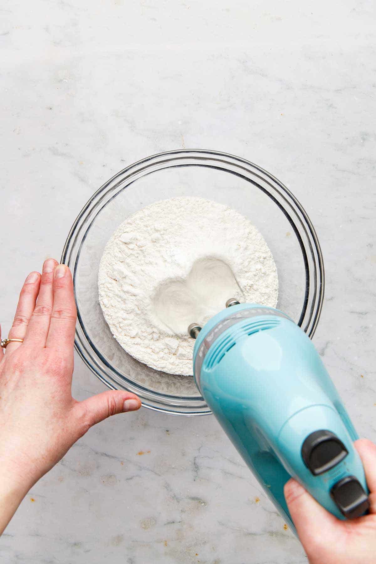 A hand using a hand mixer to mix dry cookie ingredients together in a glass bowl.