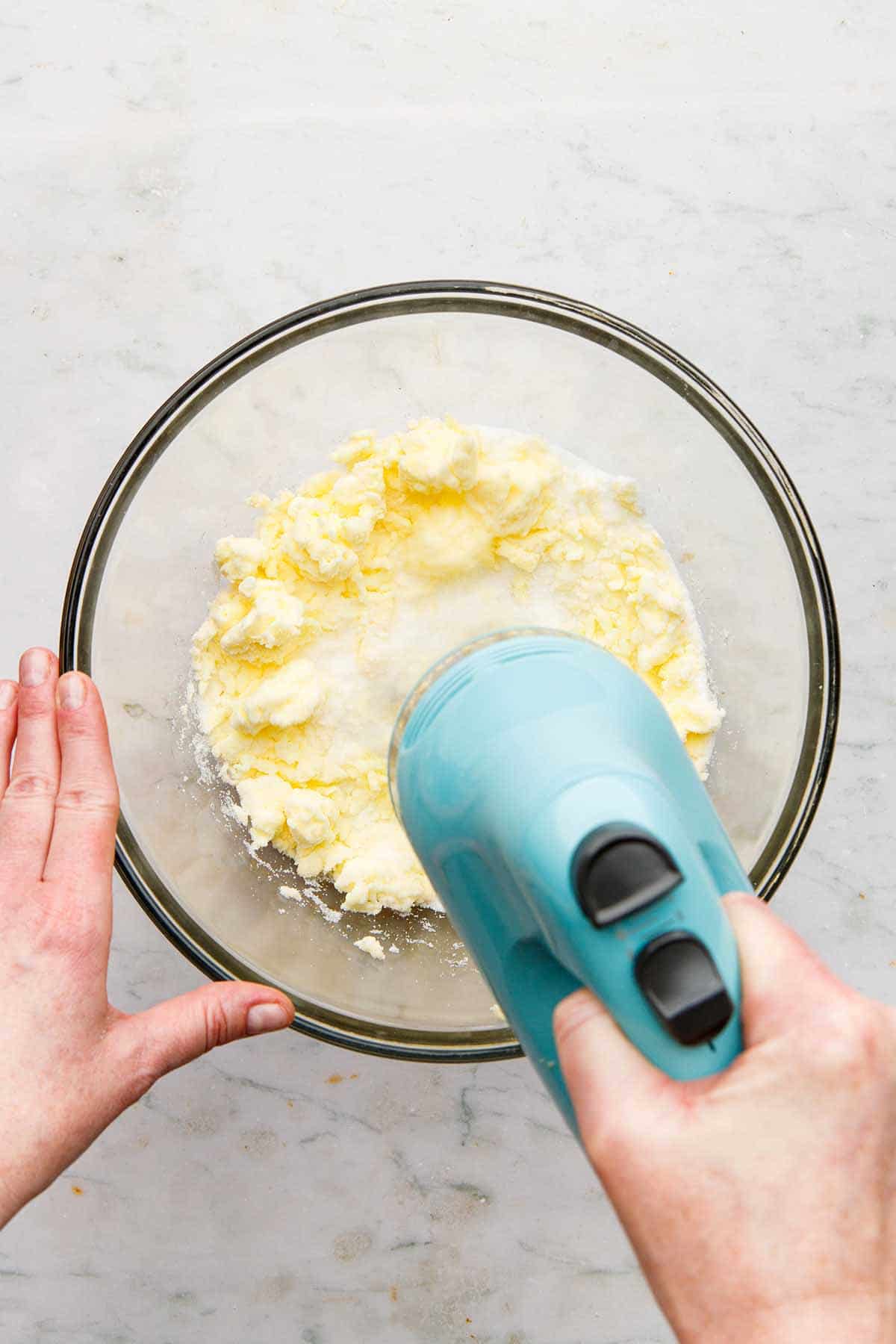 A hand using a hand mixer to beat butter and sugar in a large glass bowl.