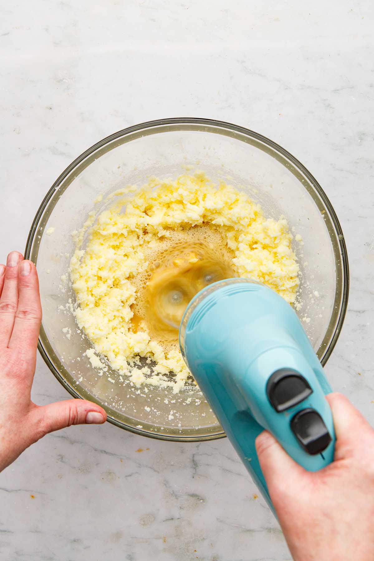 A hand using a hand mixer to mix egg mixture into wet cookie dough ingredients.