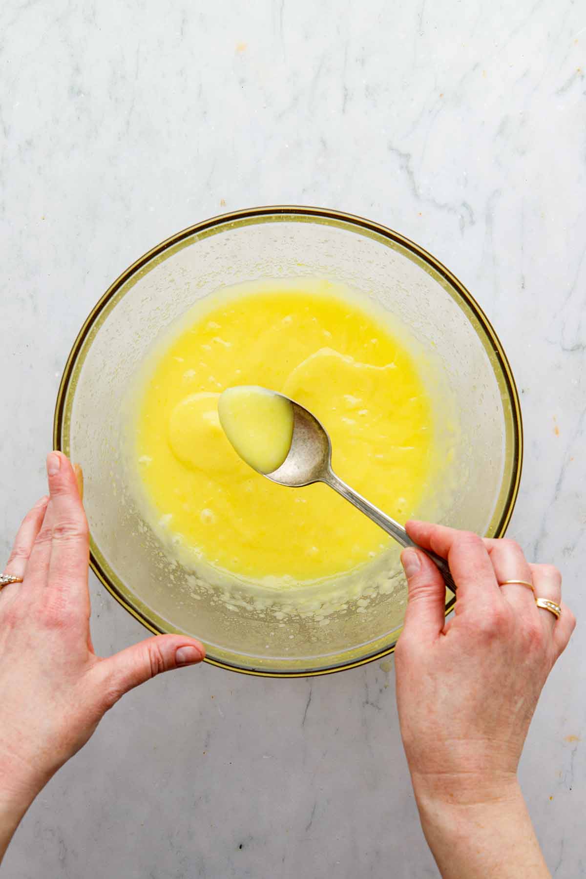 A hand holding a spoonful of curd over a bowl of cooked lemon curd.
