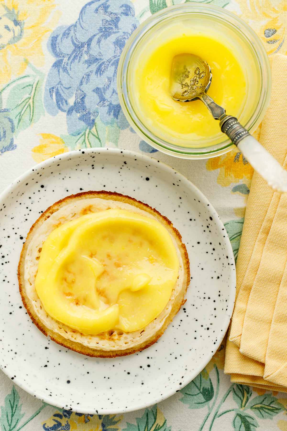 Close up overhead image of a crumpet topped with microwave lemon curd on a small white speckled plate.