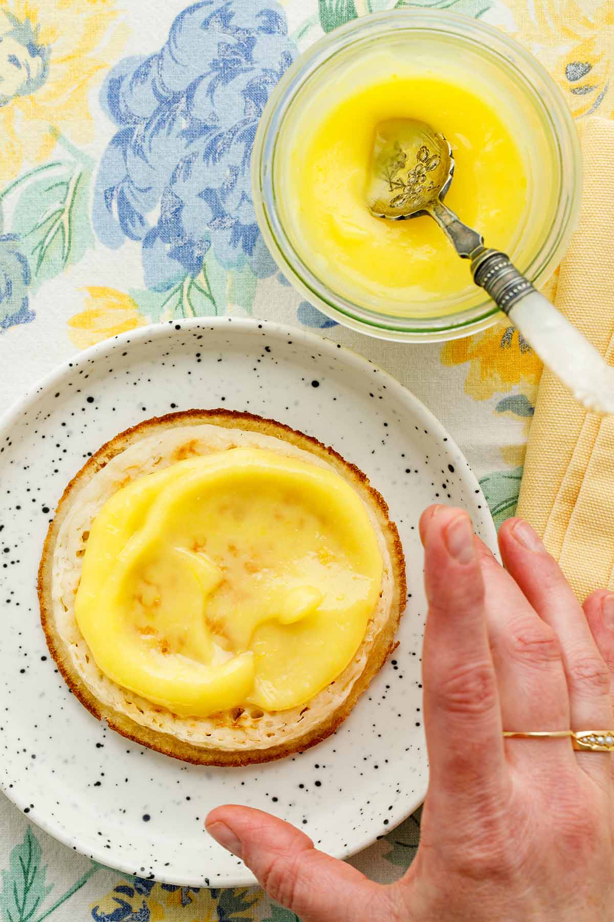 A hand reaching for a crumpet topped with microwave lemon curd on a small white speckled plate.