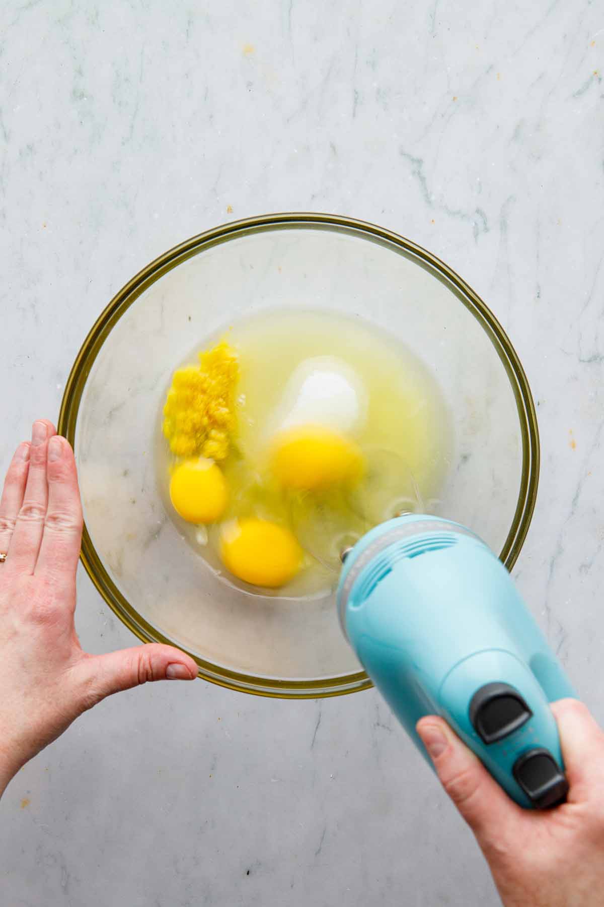 A hand using a hand mixer to whisk eggs, lemon zest, lemon juice, and sugar in a large glass bowl.