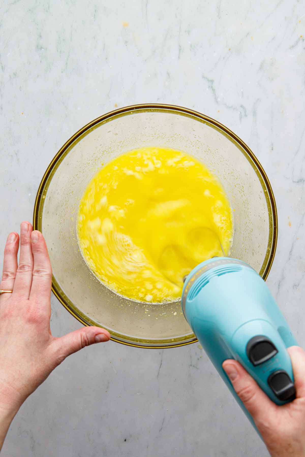 A hand using a hand mixer to mix bits of butter into a yellow liquid.