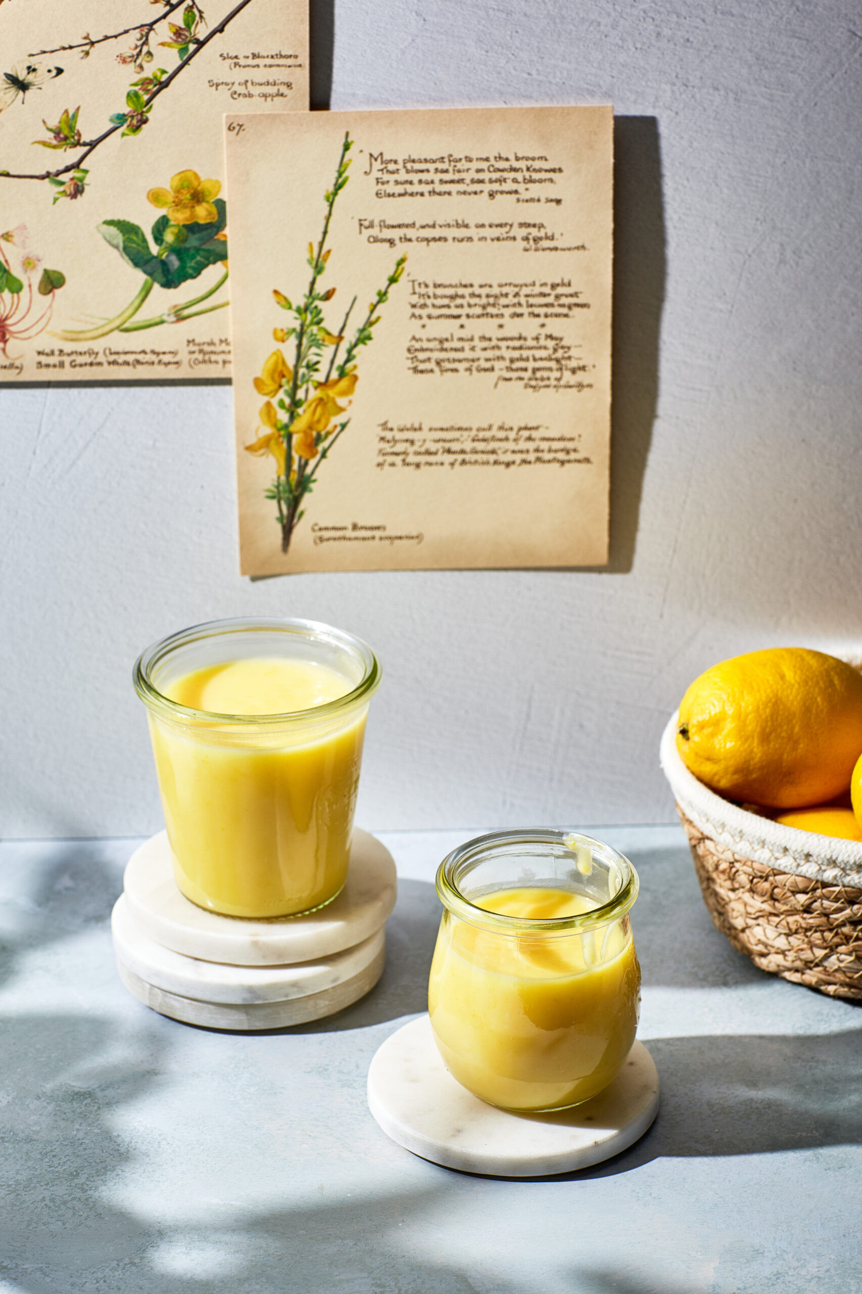 Two jars of microwave lemon curd in a sunny scene with lemons and old Edwardian book pages.