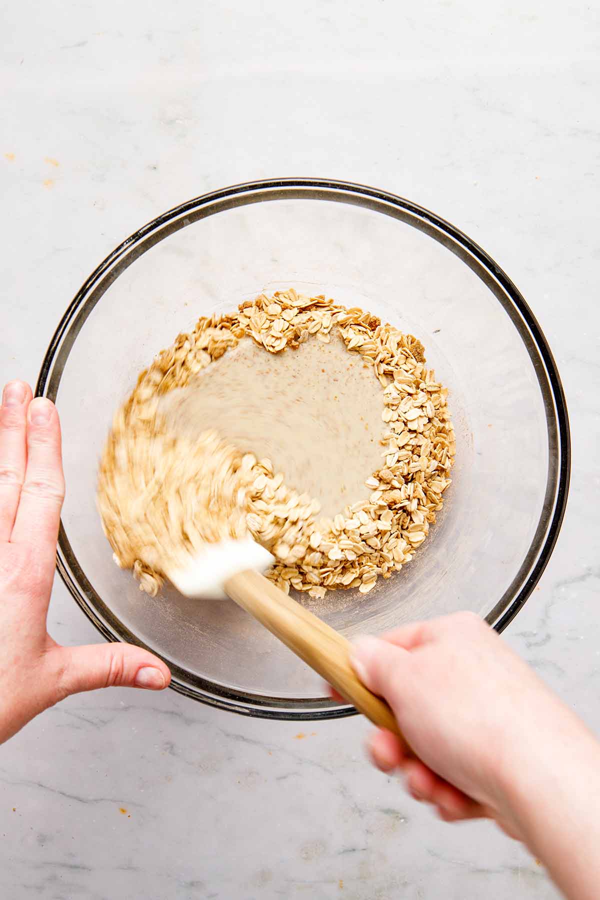 A hand using a rubber spatula to stir wet and dry ingredients in a bowl.