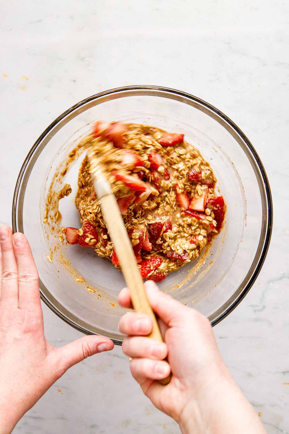 A hand stirring uncooked oatmeal and strawberries together in a bowl with a rubber spatula.