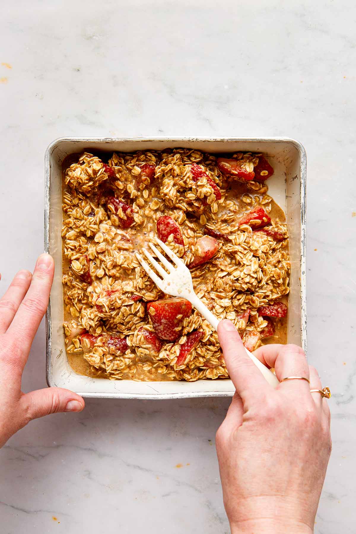 A hand using a fork to spread oatmeal and strawberries evenly in a greased baking tin.