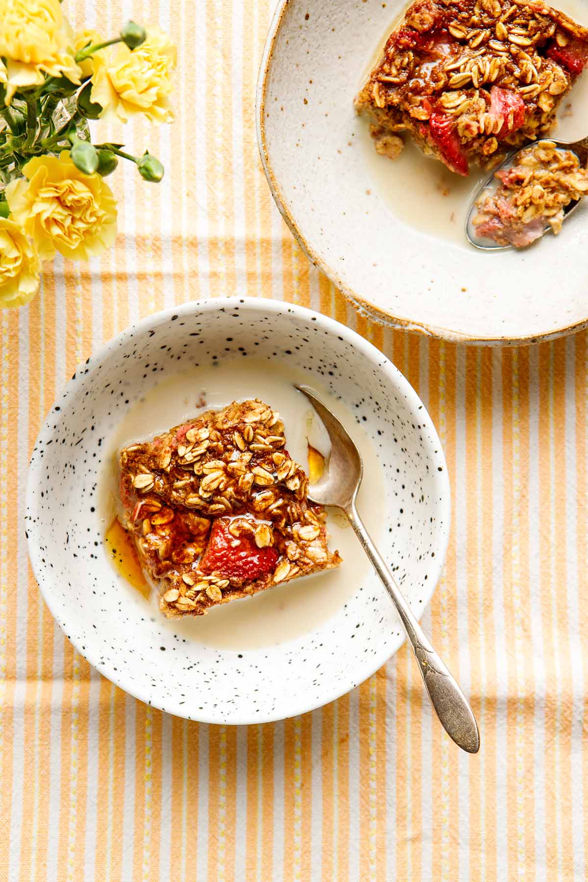 Two servings of vegan strawberry baked oatmeal in bowls on an orange striped tablecloth with a vase of yellow carnations nearby.