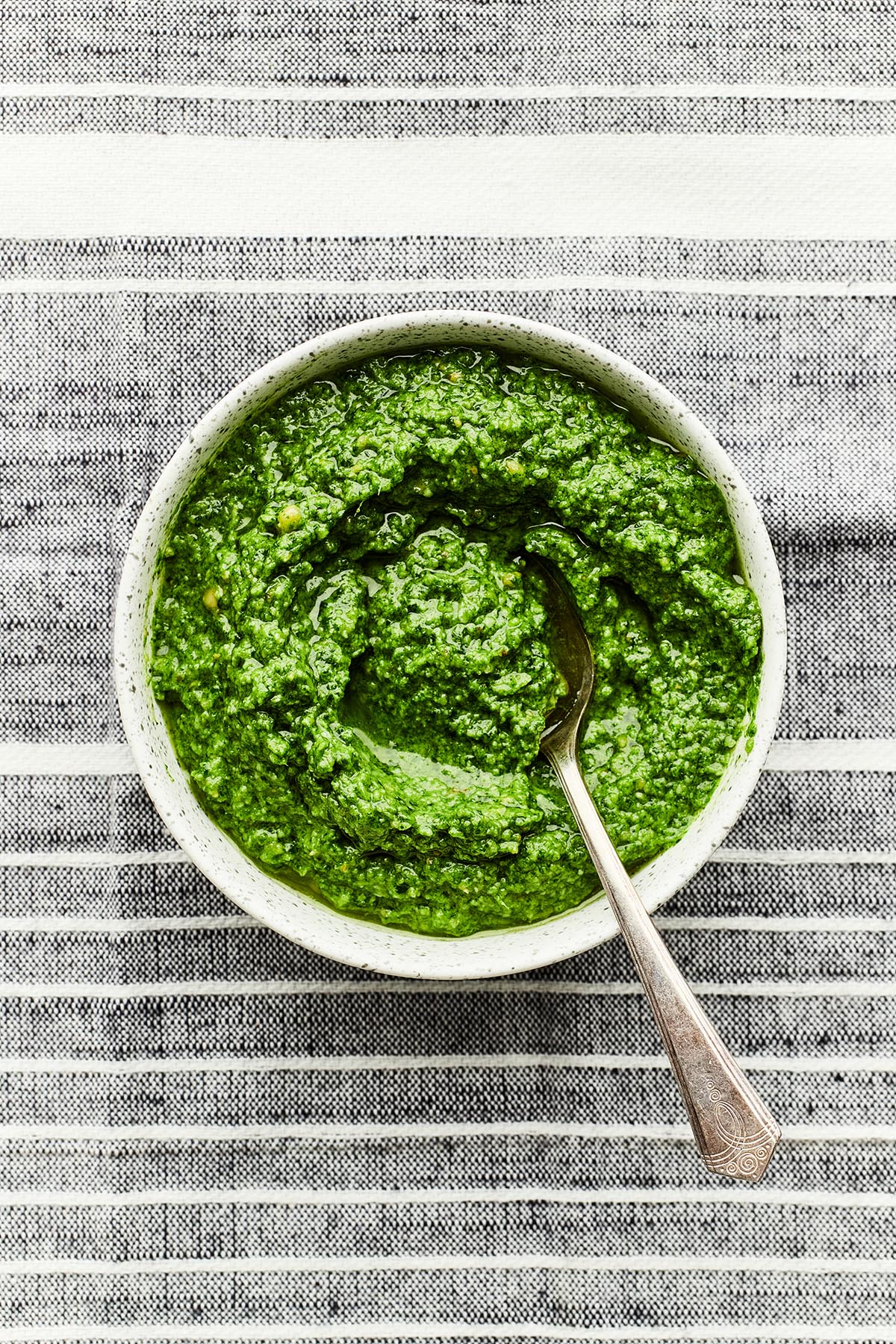 A bowl of homemade basil pesto with a spoon in the bowl on a striped tablecloth.