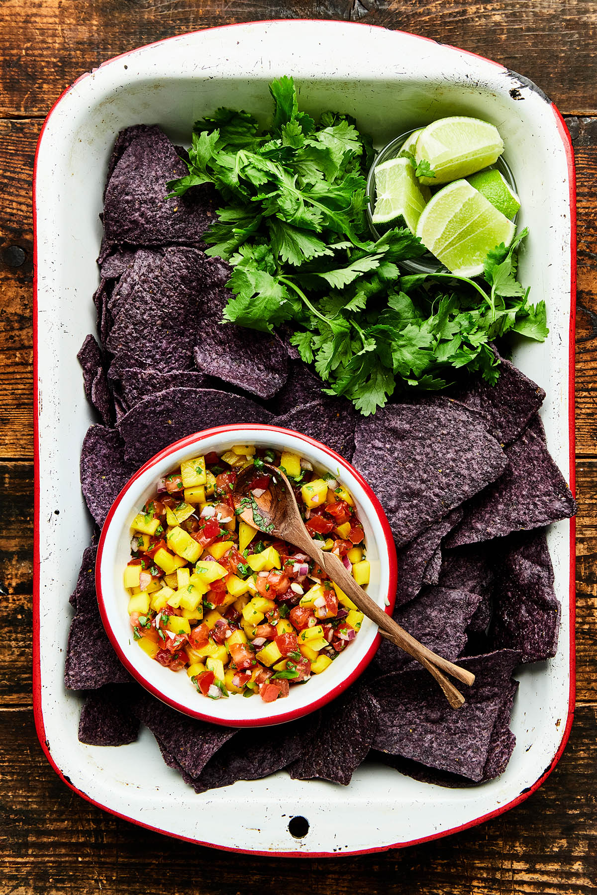 A white rectangle enamel dish with red trim filled with blue corn chips, cilantro, sliced limes, and a bowl of mango pico de gallo.