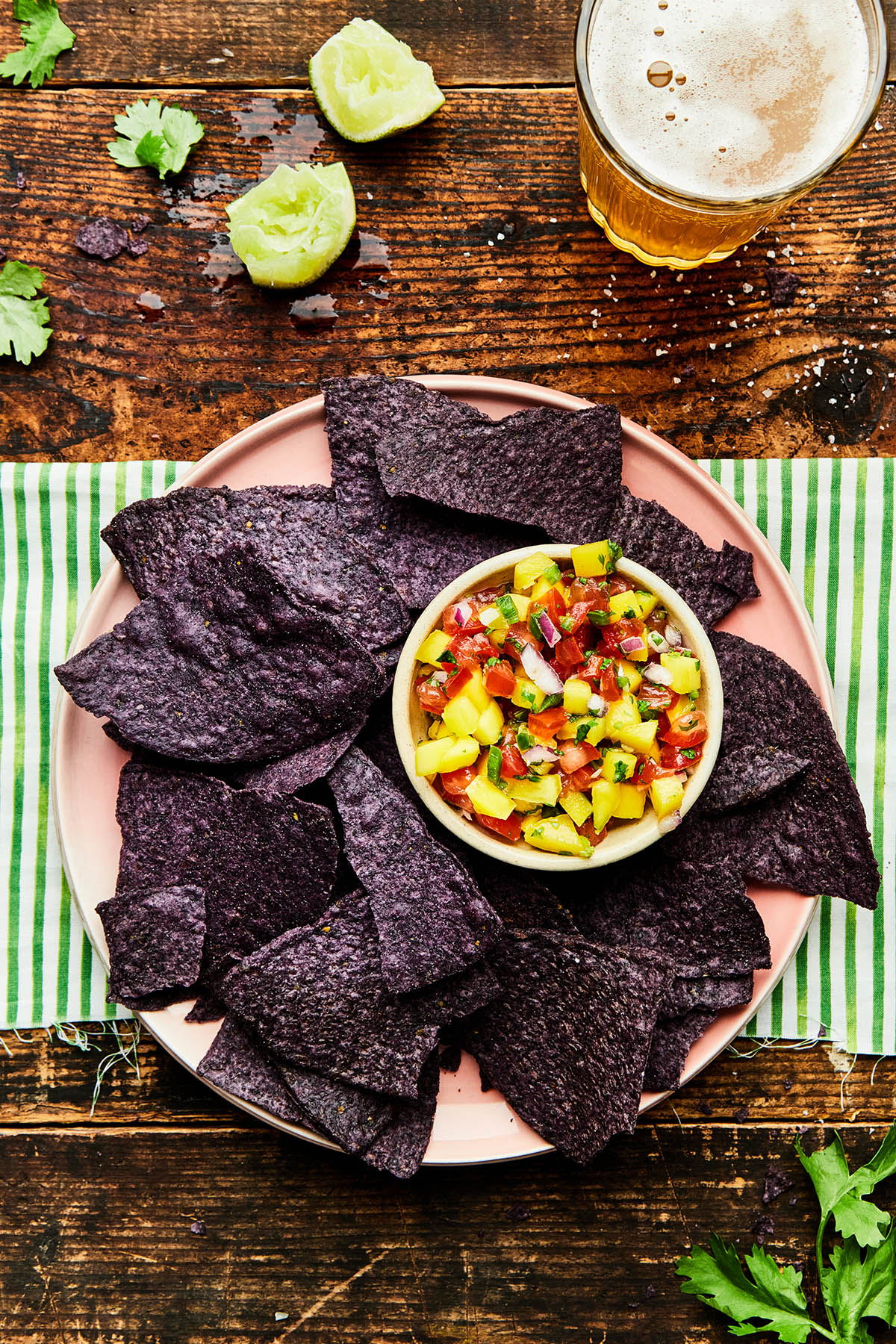 A pink plate with blue corn chips and a small bowl of mango pico de gallo laying on a green and white striped cloth on a wood table with squeezed limes, cilantro leaves, and a glass of beer nearby.