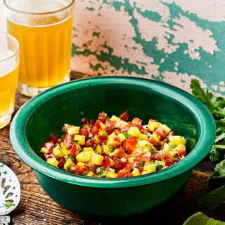 Mango pico de gallo in a green bowl on a wooden table with cilantro, sliced limes, a slotted spoon, and two glasses of beer in the background.