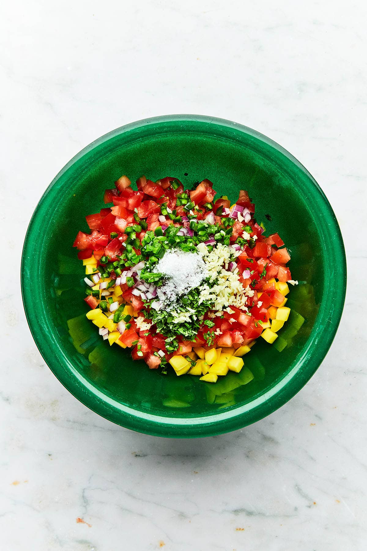 A green bowl with unmixed chopped tomatoes, mangoes, jalapeno, cilantro, red onion, garlic, and salt inside.