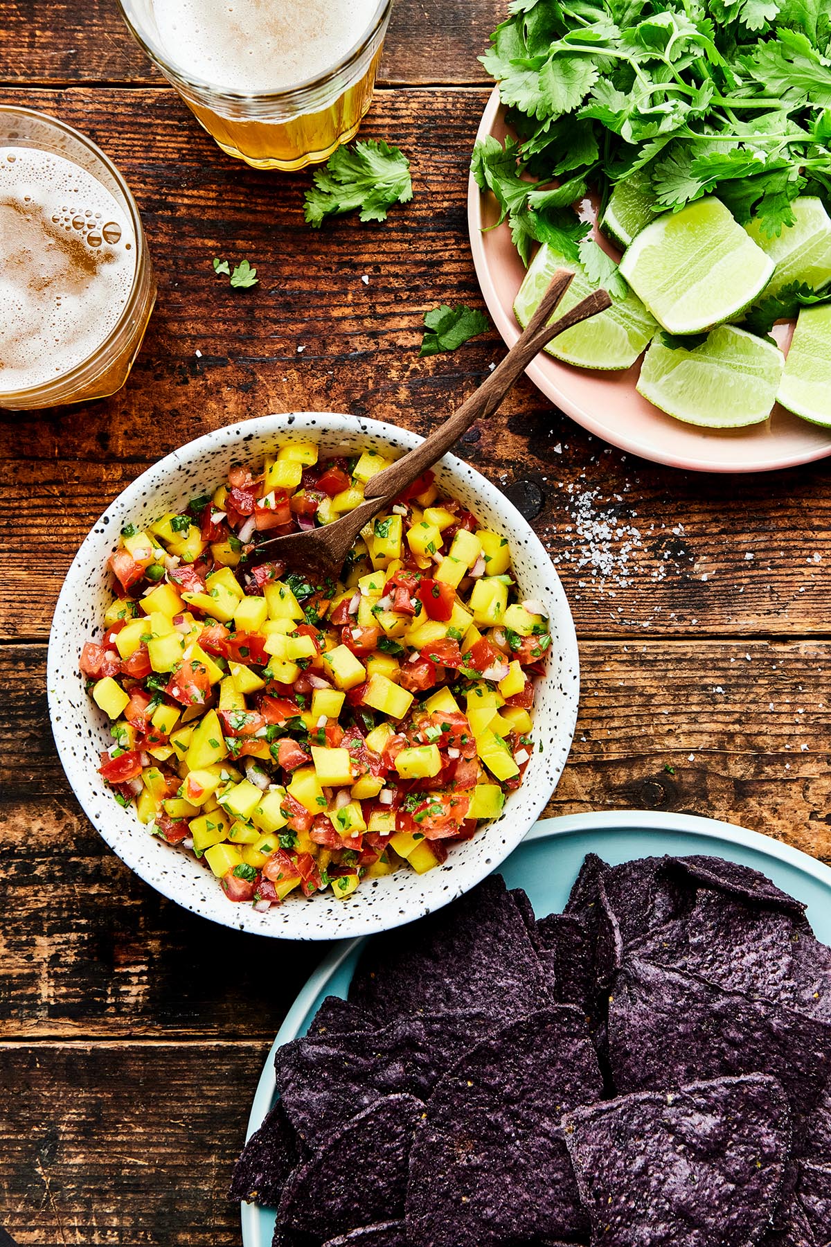 A bowl of mango pico de gallo on a wooden table alongside a blue plate with blue corn chips, a pink plate with fresh cilantro and limes, and two glasses of beer.